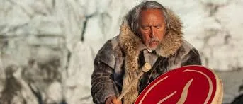 An elder with long grey hair in a fur-trimmed parka beats a red hand drum in front of a wall of glacial ice.