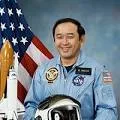 Official portrait of astronaut Ellison Onizuka in a blue flight suit, with an American flag and a space shuttle model behind him.