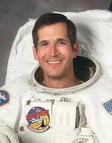Smiling astronaut John Herrington in a white spacesuit with mission patches, his helmet removed.