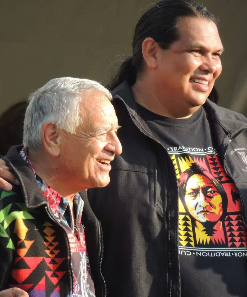 Rueben George, a younger man with long dark hair in a T-shirt bearing a portrait of a Native chief, smiles with his arm around a white-haired elder in a geometric-patterned jacket.