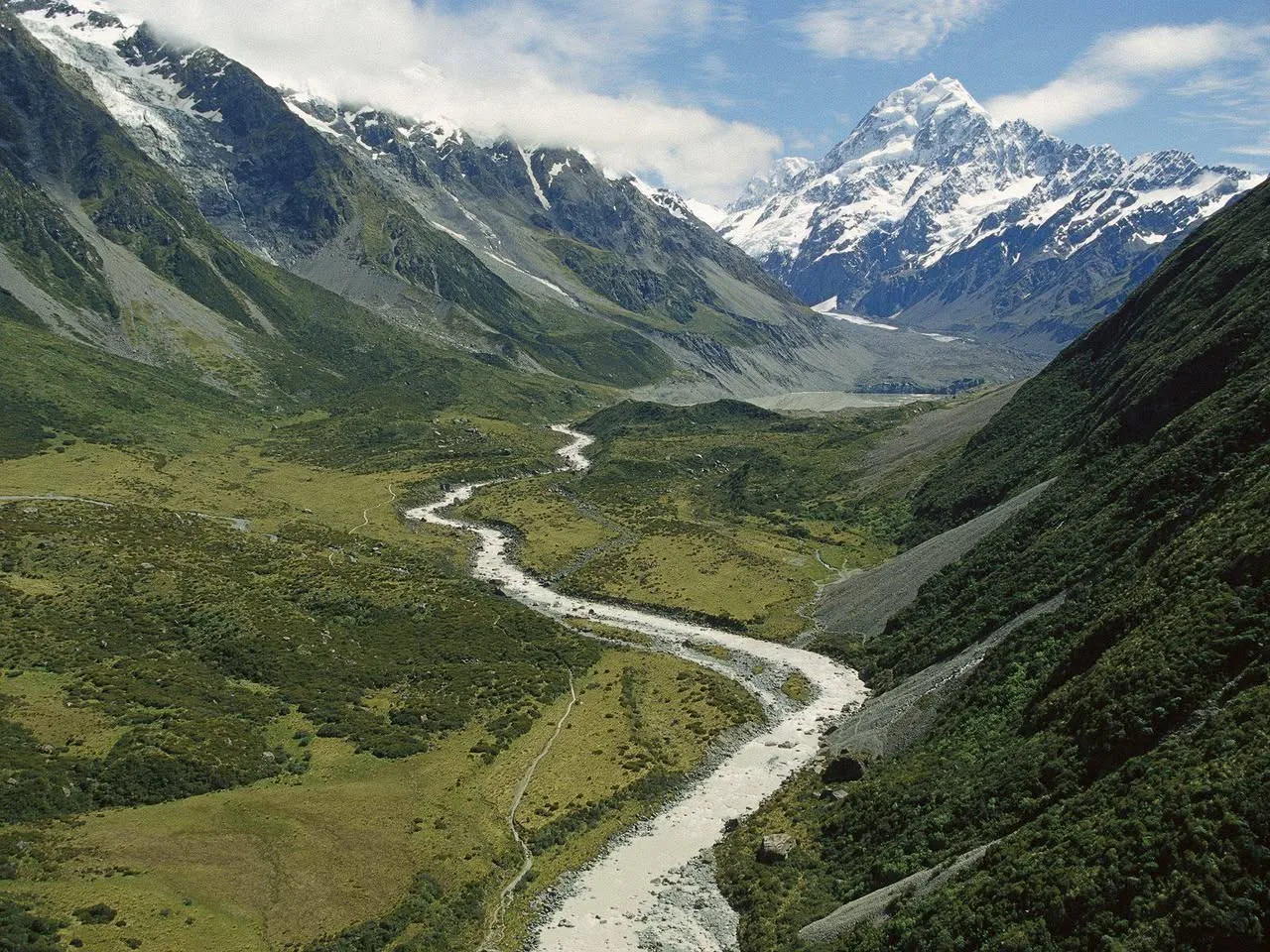 A glacial river winds through a green valley between steep mountain slopes toward a snow-capped peak under a blue sky with drifting clouds.