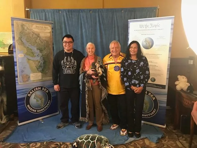 Four people stand together on a blue cloth between Salish Sea Marine Sanctuary banners, one showing a map of the Salish Sea and one a 'We the People' proclamation.
