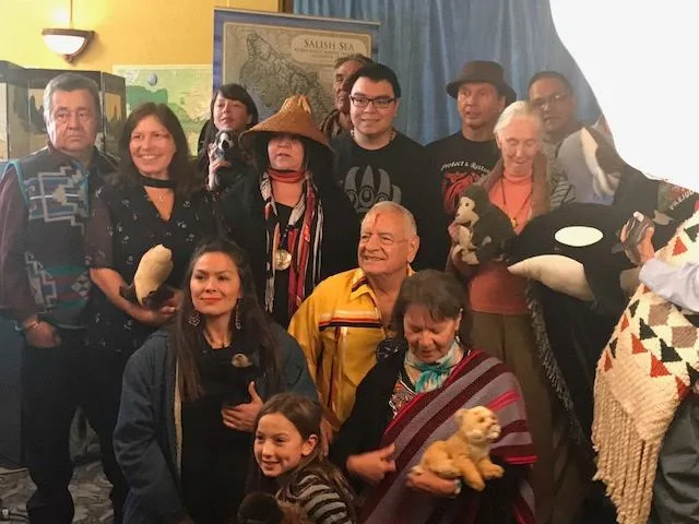 A large group of Indigenous elders, adults and a child gather in front of a Salish Sea map, several holding stuffed animals including an orca and a chimpanzee.
