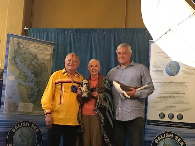 An elder in a yellow ribbon shirt, a white-haired woman holding a stuffed chimpanzee, and a man holding a stuffed orca stand between Salish Sea Marine Sanctuary banners.