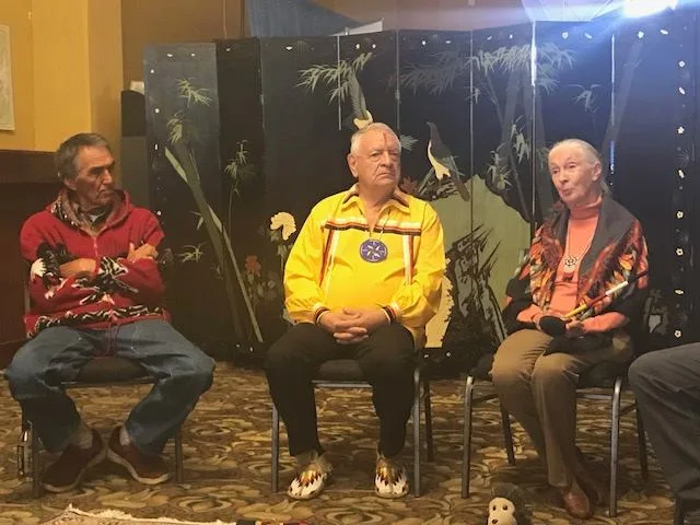 Three elders seated before a painted folding screen: a man in a red patterned sweater, a man in a yellow ribbon shirt, and a white-haired woman speaking.