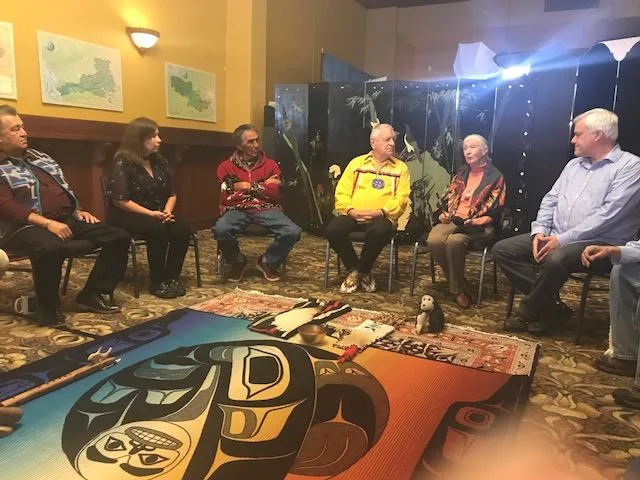 A circle of people seated around a Northwest Coast design blanket on the floor, with maps of the Salish Sea on the yellow wall behind them.