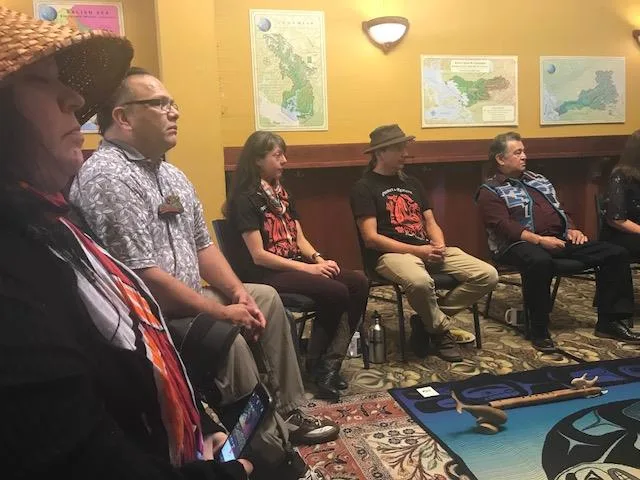 Participants seated in a row listening attentively, one in a woven cedar hat in the foreground, maps of the Salish Sea region on the wall.