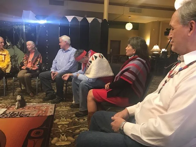 Side view of the circle, with a man in a white shirt in the foreground and others in traditional regalia and blankets listening to a white-haired woman.
