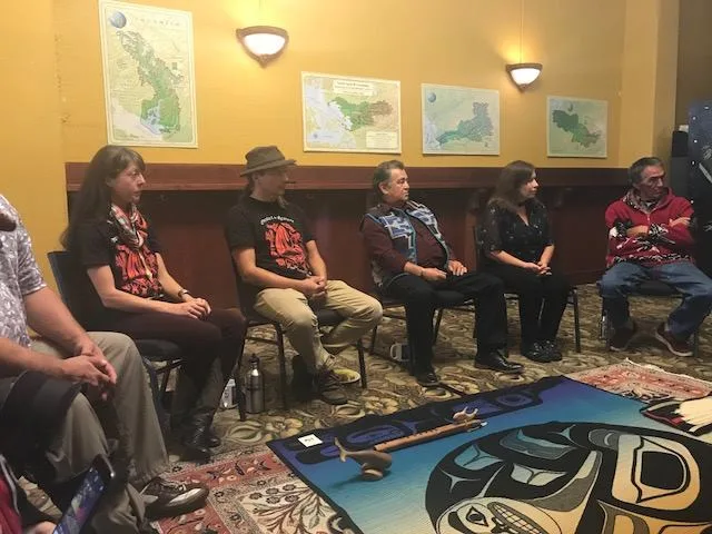 People seated in a row against a yellow wall hung with regional maps, facing a Northwest Coast design blanket with ceremonial items on it.