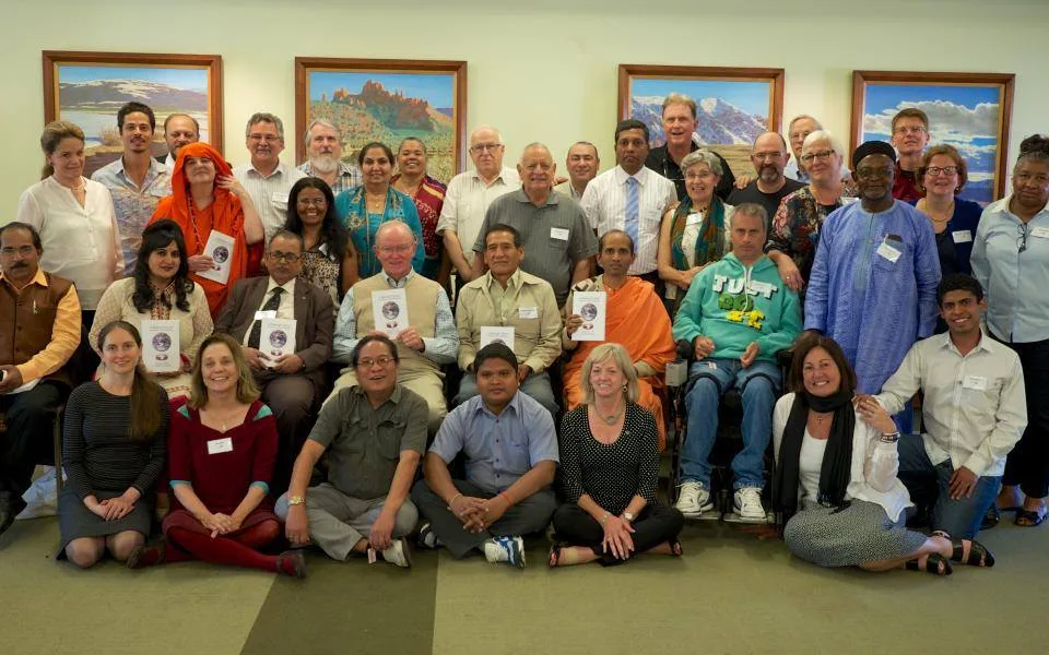 Large group portrait of URI trustees, staff and council members of diverse faiths and cultures, some holding booklets with an Earth image, posed before landscape paintings.