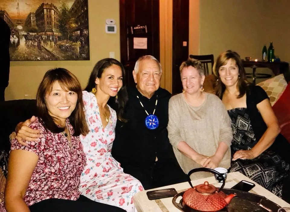 Phil Lane Jr., wearing a blue beaded star medallion, sits smiling on a couch with four women in a warmly lit room, a red teapot on the table before them.
