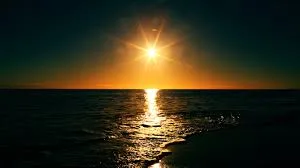 The sun shines low over a calm sea, casting a golden path of light across the dark water beneath a deepening sky.