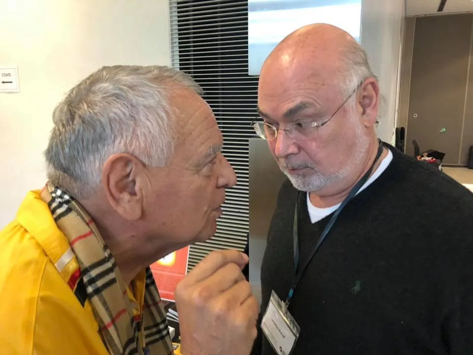 Phil Lane Jr. and Jonathan Granoff in close conversation: a grey-haired man in a yellow shirt and plaid scarf speaks to a bald man with glasses, a dark sweater and a name badge.