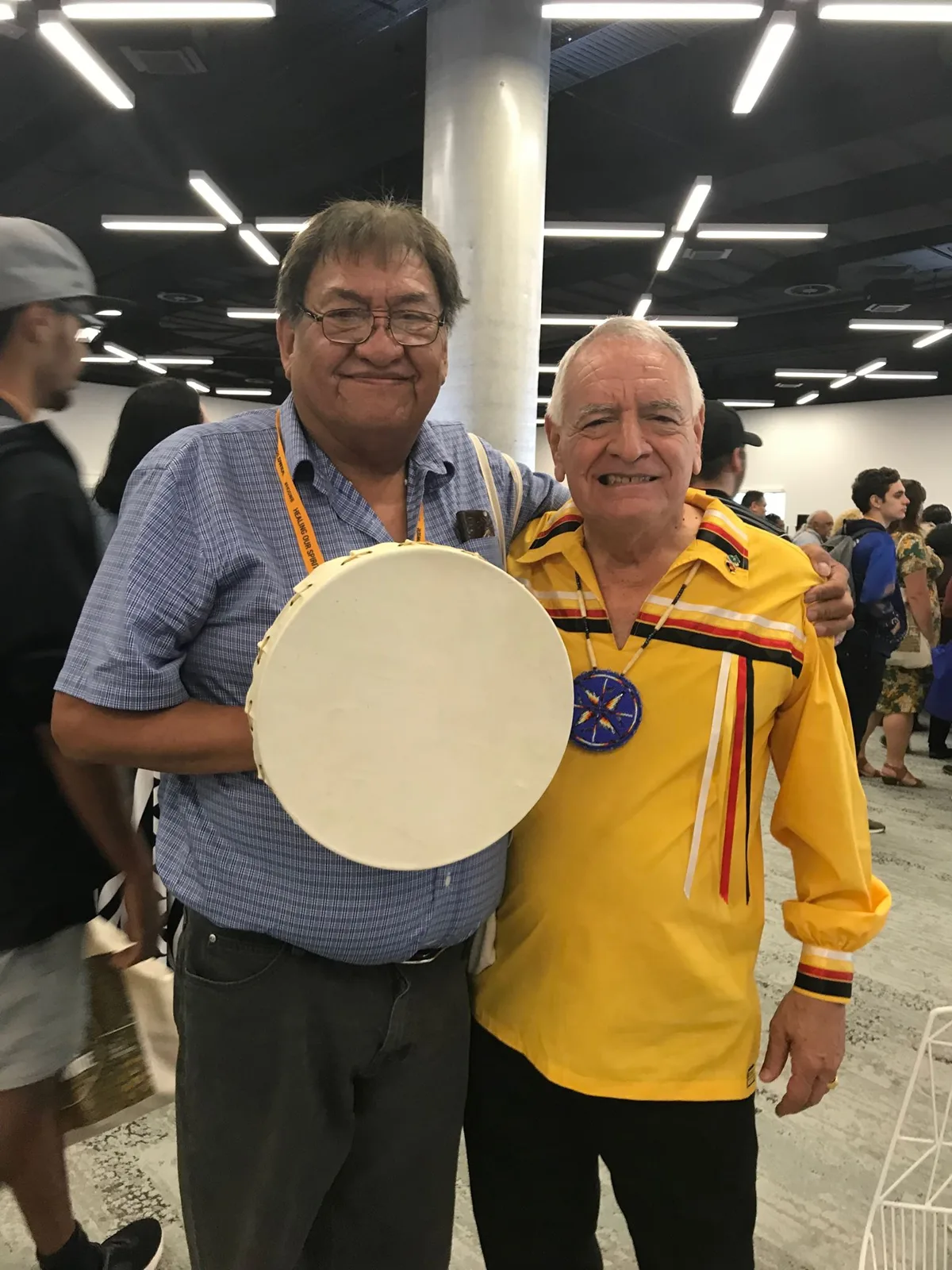 Old friends Freddie Johnson and Phil Lane Jr. smile arm in arm in a busy conference hall; one holds a hand drum, the other wears a yellow ribbon shirt and beaded medallion.