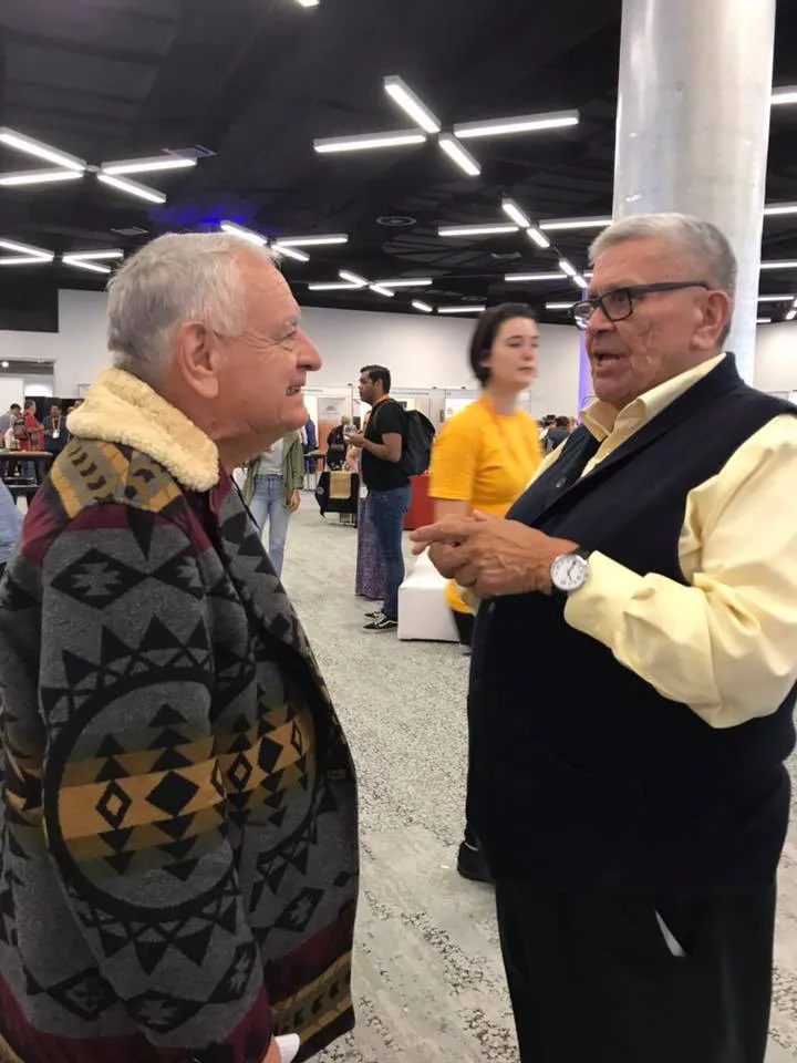 Two elderly men talk warmly in a busy conference hall; one wears a patterned blanket coat with a fleece collar, the other a yellow shirt, dark vest and glasses.