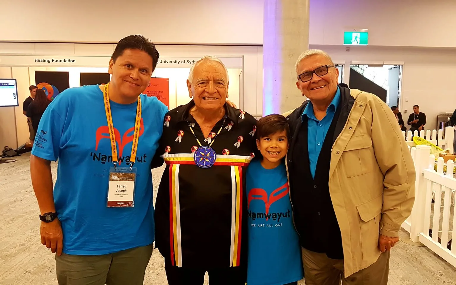 Farrell Joseph, wearing a conference badge, and a smiling boy, both in blue 'Namwayut We Are All One' shirts, stand with an elder in a ribbon shirt and an elder in glasses.