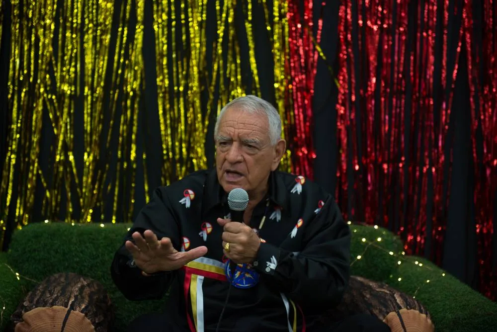 A white-haired elder in a black ribbon shirt with medicine wheel and feather designs speaks into a microphone, gesturing, before gold and red tinsel curtains.