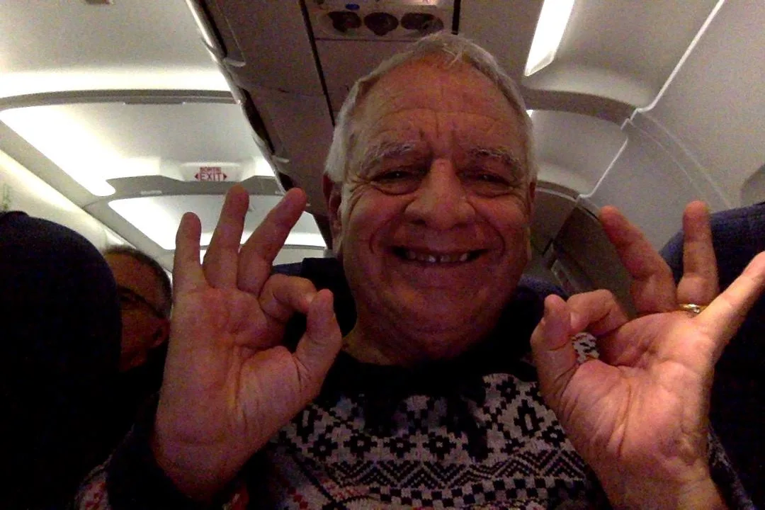 Phil Lane Jr., a smiling older man with grey hair in a patterned sweater, takes a selfie aboard an airplane, making OK signs with both hands.