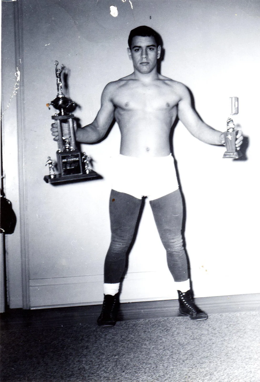 Black-and-white photo of a young Phil Lane Jr., age seventeen, shirtless in wrestling trunks, tights and boots, holding a large trophy in one hand and a smaller trophy in the other.