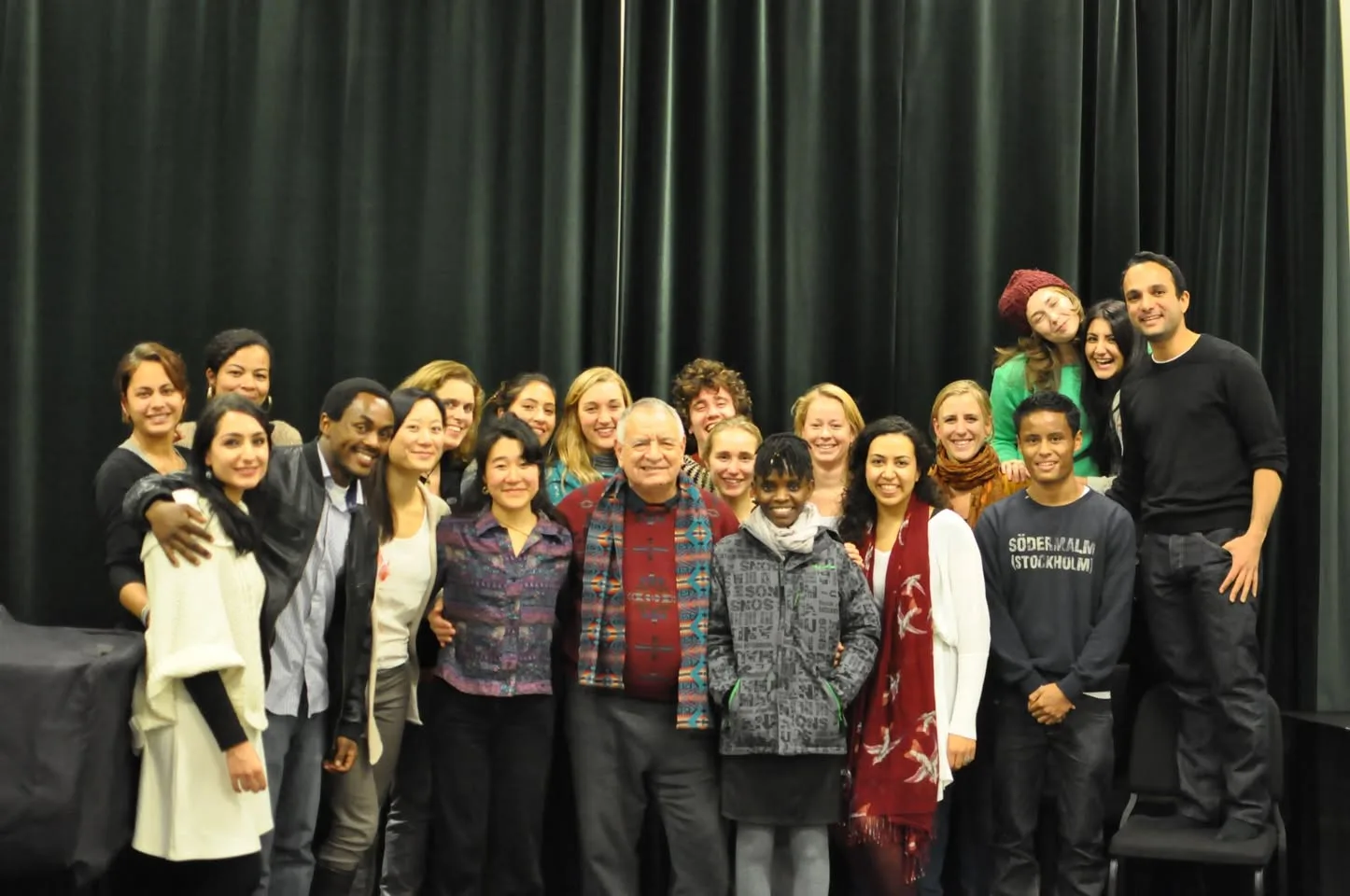 A large, smiling group of young adults from many backgrounds poses before a dark green curtain, gathered around an older man in a red sweater and patterned scarf.