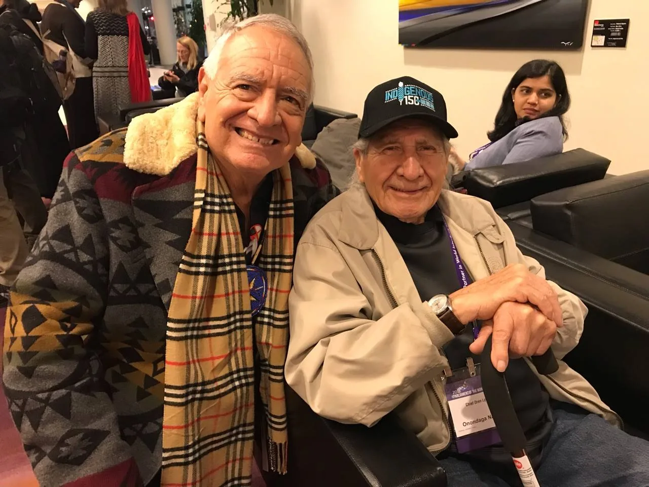 Phil Lane Jr., smiling in a patterned jacket and plaid scarf, sits beside Chief Oren Lyons, who wears a black Indigenous cap, a conference badge and rests his hands on a cane.