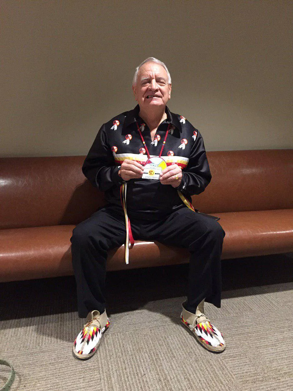 Phil Lane Jr., a white-haired elder in a black ribbon shirt with feather designs and beaded moccasins, sits on a leather bench holding up his Ihanktonwan Nation passport card.