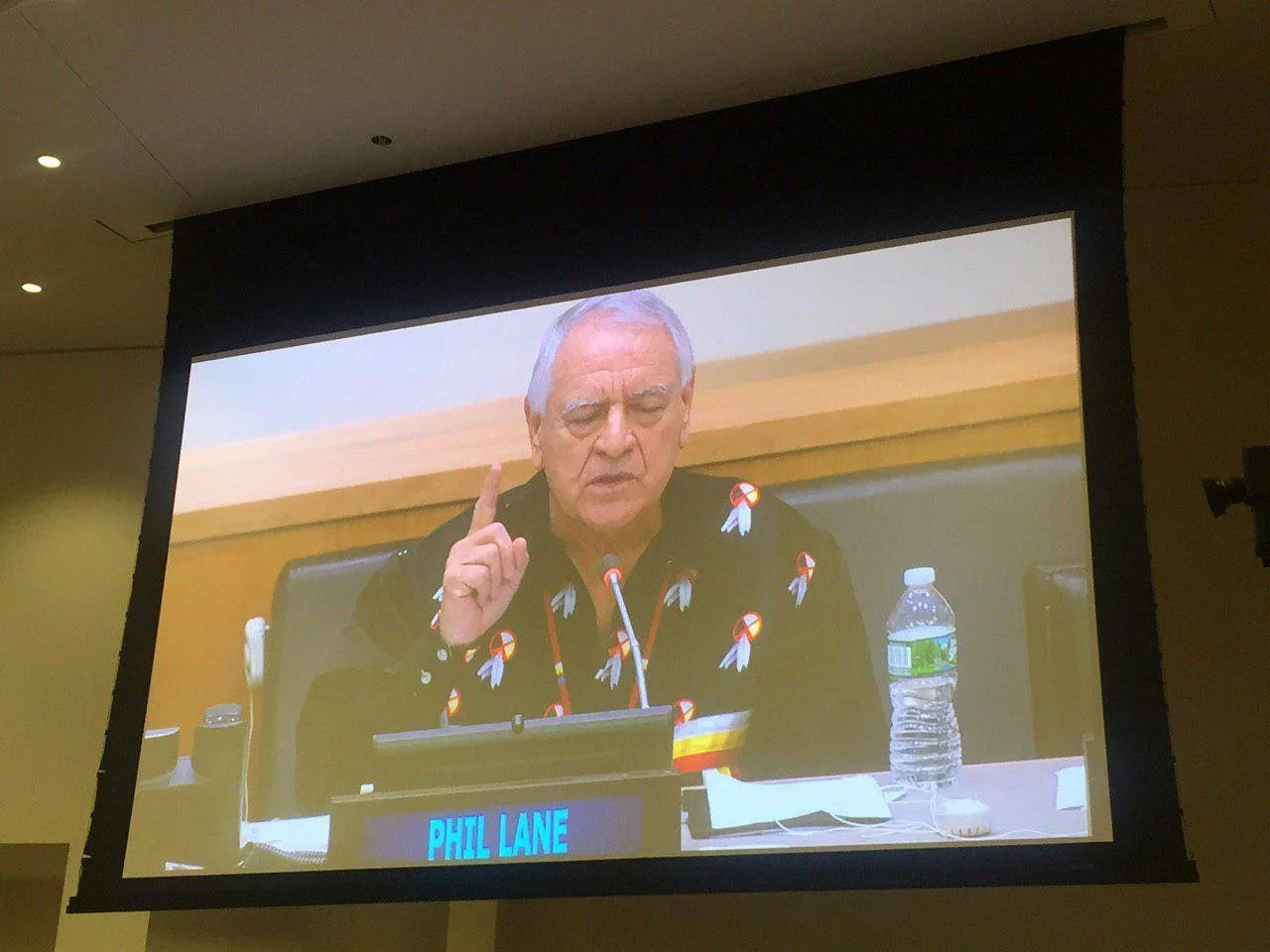 A projection screen showing Phil Lane, with grey hair and a black shirt decorated with medicine wheel and feather designs, raising a finger as he speaks behind a nameplate.