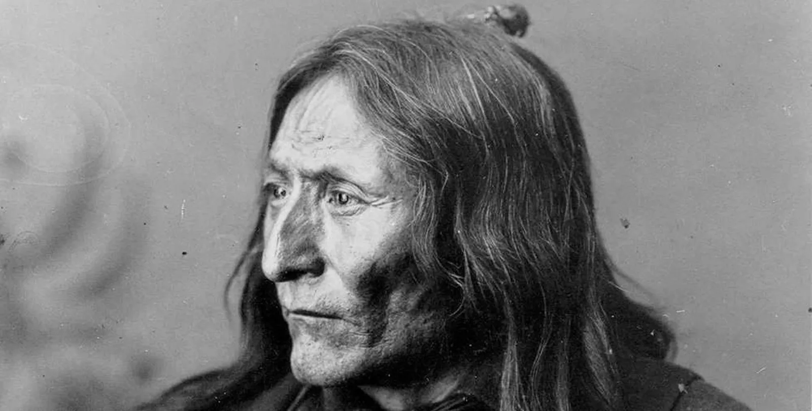 Black-and-white historical portrait of Chief Crowfoot, an elder with long hair and a deeply lined face, gazing off to the side.