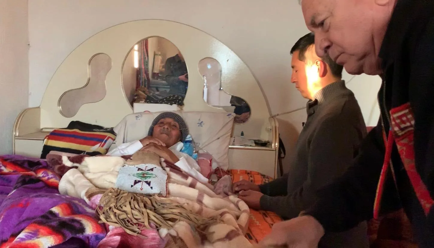 An elderly man in a knit cap rests in bed under blankets and a beaded fringed bundle, while a young man and an older man in a woven red sash lean in beside him in prayer.