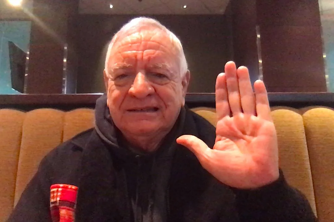 An elderly man with white hair, wearing a dark hooded jacket, raises his open right hand in greeting while seated in a booth with tan upholstered seating.