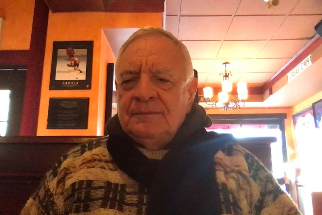 An older man with white hair, wearing a dark scarf and patterned sweater, sits in a restaurant with orange walls, framed pictures and a hanging chandelier.