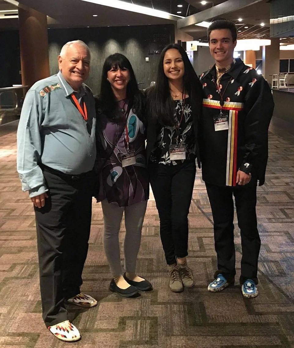 Phil Lane Jr., his grandson Hudson, Lisa Paz and Danielle Arpan stand smiling in a conference venue hallway, wearing name badges; Phil and Hudson wear beaded moccasins and Hudson a ribbon shirt.