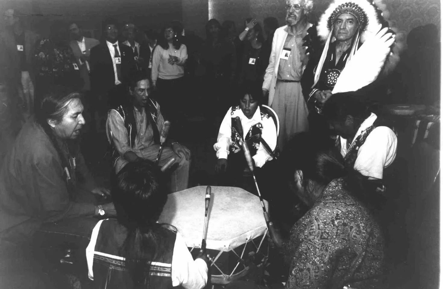 Black-and-white photo of singers seated around a large hand drum, beating it with drumsticks, as a crowd with name badges and an elder in a feathered headdress look on.