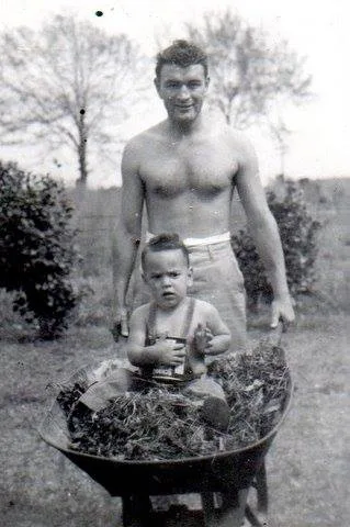 A black-and-white photo of a shirtless young man pushing a wheelbarrow with a toddler sitting in it outdoors.