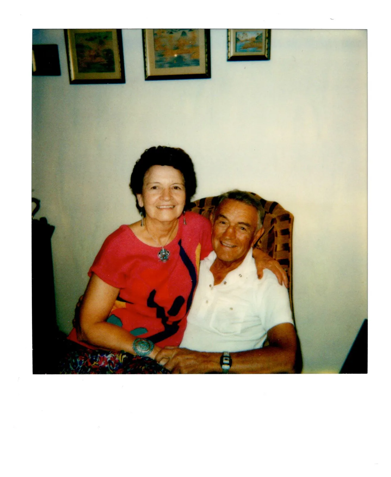 A smiling woman in a red top and turquoise jewelry with her arm around Mato Gi, seated in a white shirt in an armchair.