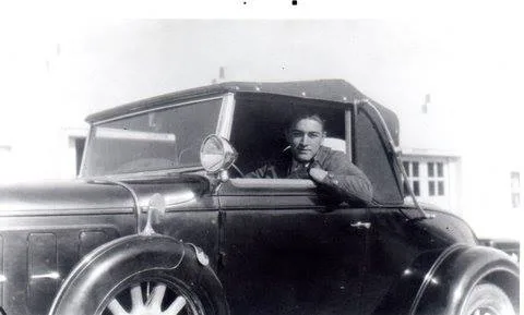 A black-and-white photo of a young man looking out from the driver's seat of a vintage convertible car.