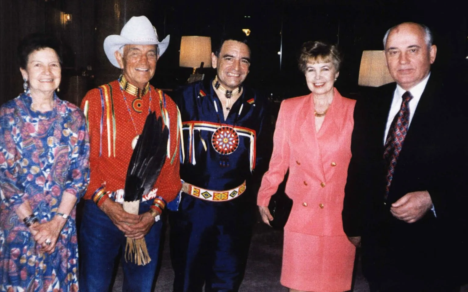 Five people smiling indoors: an older woman in a floral dress, an older man in a cowboy hat and red ribbon shirt holding an eagle feather fan, Phil Lane Jr. in a blue ribbon shirt, Raisa Gorbachev in a pink suit, and Mikhail Gorbachev.