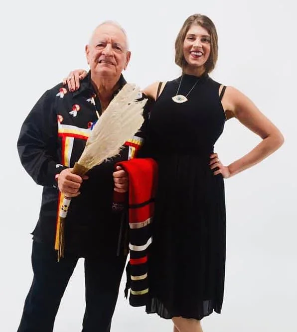 Phil Lane Jr., in a black ribbon shirt and holding a white feather fan and a folded red blanket, stands arm in arm with Tonja Toni Lane, smiling in a black dress, against a white backdrop.
