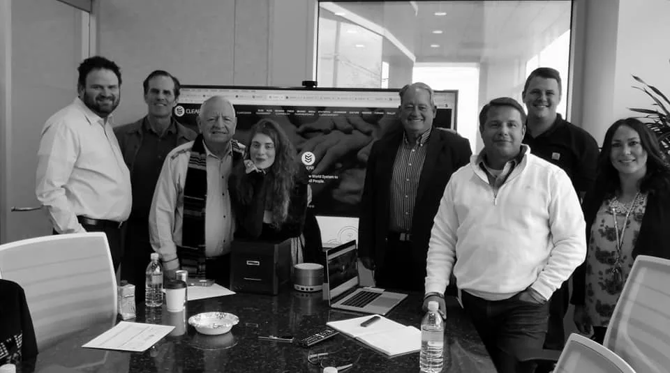 Black-and-white group photo of nine people standing around a conference table in front of a large screen displaying a Clear website.