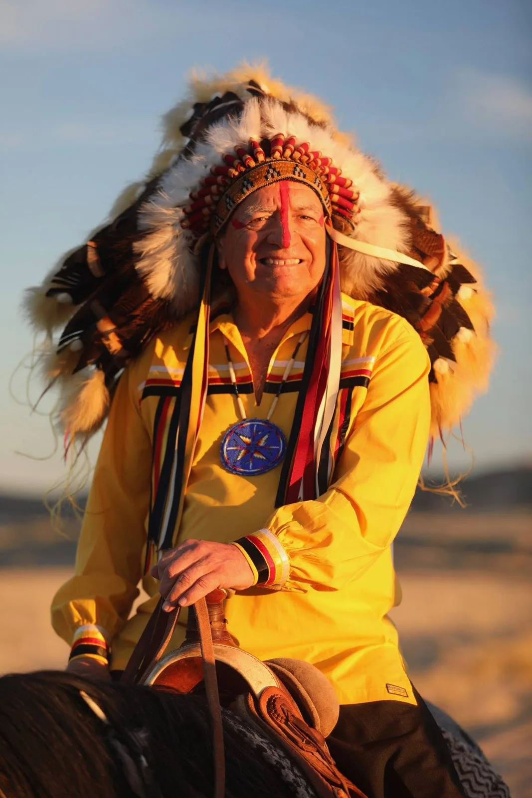 Phil Lane Jr. smiling on horseback at sunset, wearing a feathered headdress, red face paint, a yellow ribbon shirt and a blue beaded medallion.