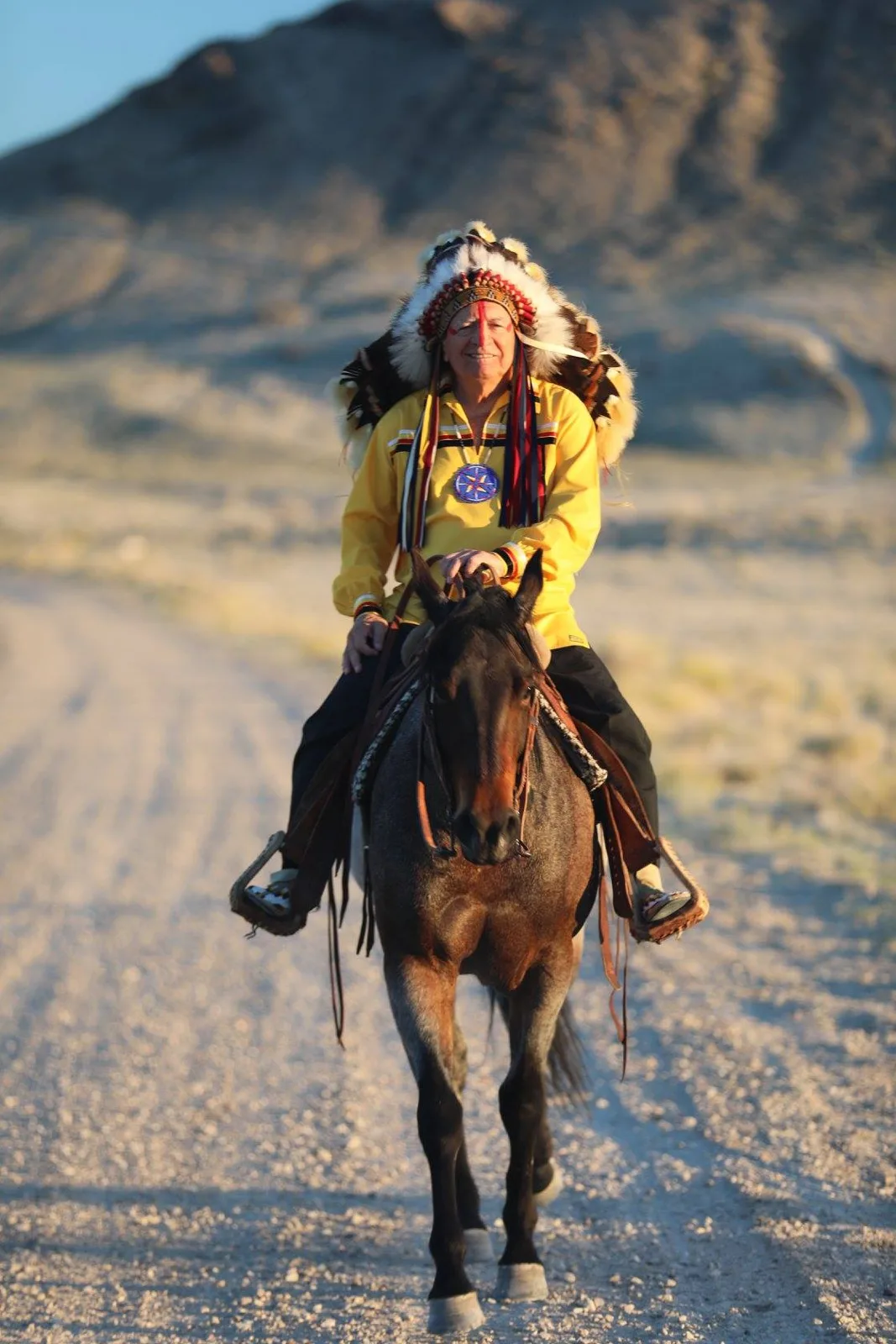 Phil Lane Jr. in a feathered headdress and yellow ribbon shirt riding a dark horse toward the camera along a gravel road in the desert.