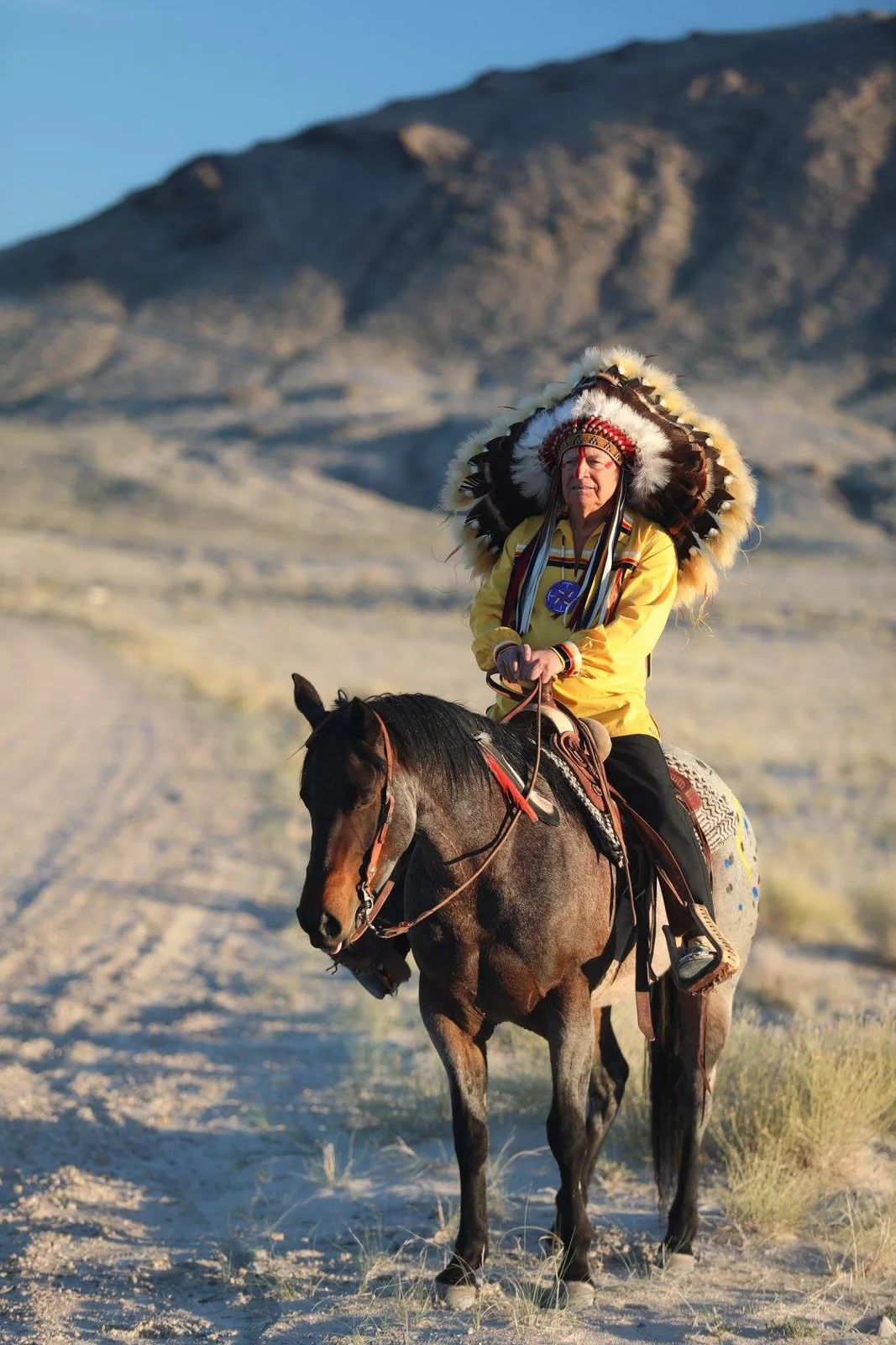 Phil Lane Jr. in a feathered headdress and yellow ribbon shirt seated on a roan horse beside a dirt road, with a rocky desert hill behind.