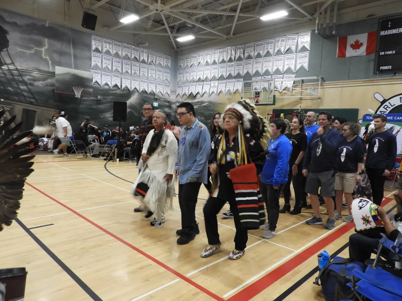A grand entry procession in a school gymnasium led by an elder in a feather headdress, a man in white fringed buckskin, and a young man in a blue shirt, followed by supporters in matching sweatshirts.