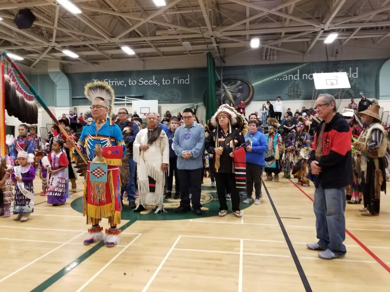 Dancers in colorful regalia, elders, and a young man in a blue shirt stand with heads bowed in a gymnasium during a powwow ceremony, beneath a wall reading "To Strive, to Seek, to Find."