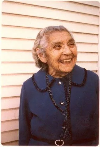 Grandmother Martina Sherwood, an elderly woman with gray hair, smiling broadly in a navy dress with a dark necklace, standing before white wooden siding.