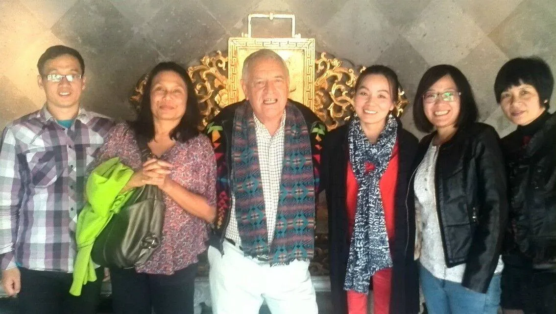 An elder with white hair wearing a patterned woven scarf stands smiling with five companions in front of an ornate gold frame on a stone wall.