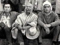 Black-and-white photograph of three men seated side by side outdoors; the man in the middle wears a striped poncho and knit cap and holds a wide-brimmed hat on his knee.