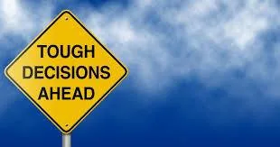A yellow diamond-shaped road sign reading "Tough Decisions Ahead" against a deep blue sky with white clouds.