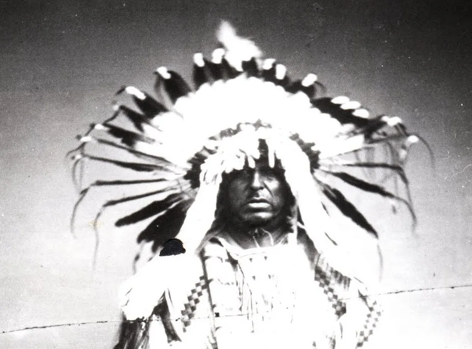 Black-and-white archival portrait of a Lakota man in a full eagle-feather war bonnet and beaded regalia, gazing solemnly at the camera.