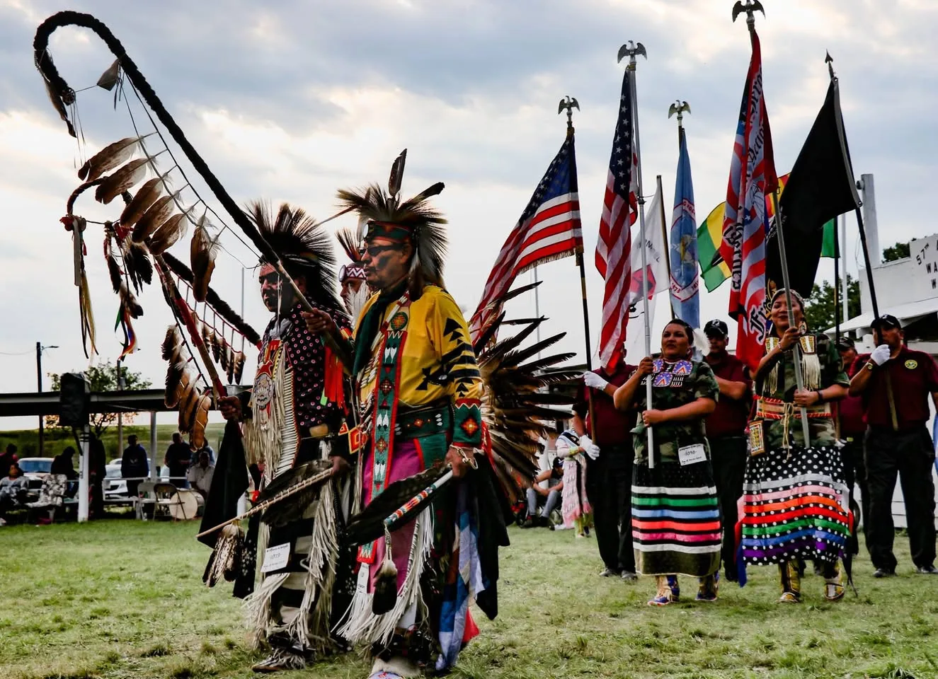 Two men in feathered regalia with bustles lead a grand entry at a powwow, followed by flag bearers carrying the U.S. flag and other flags.